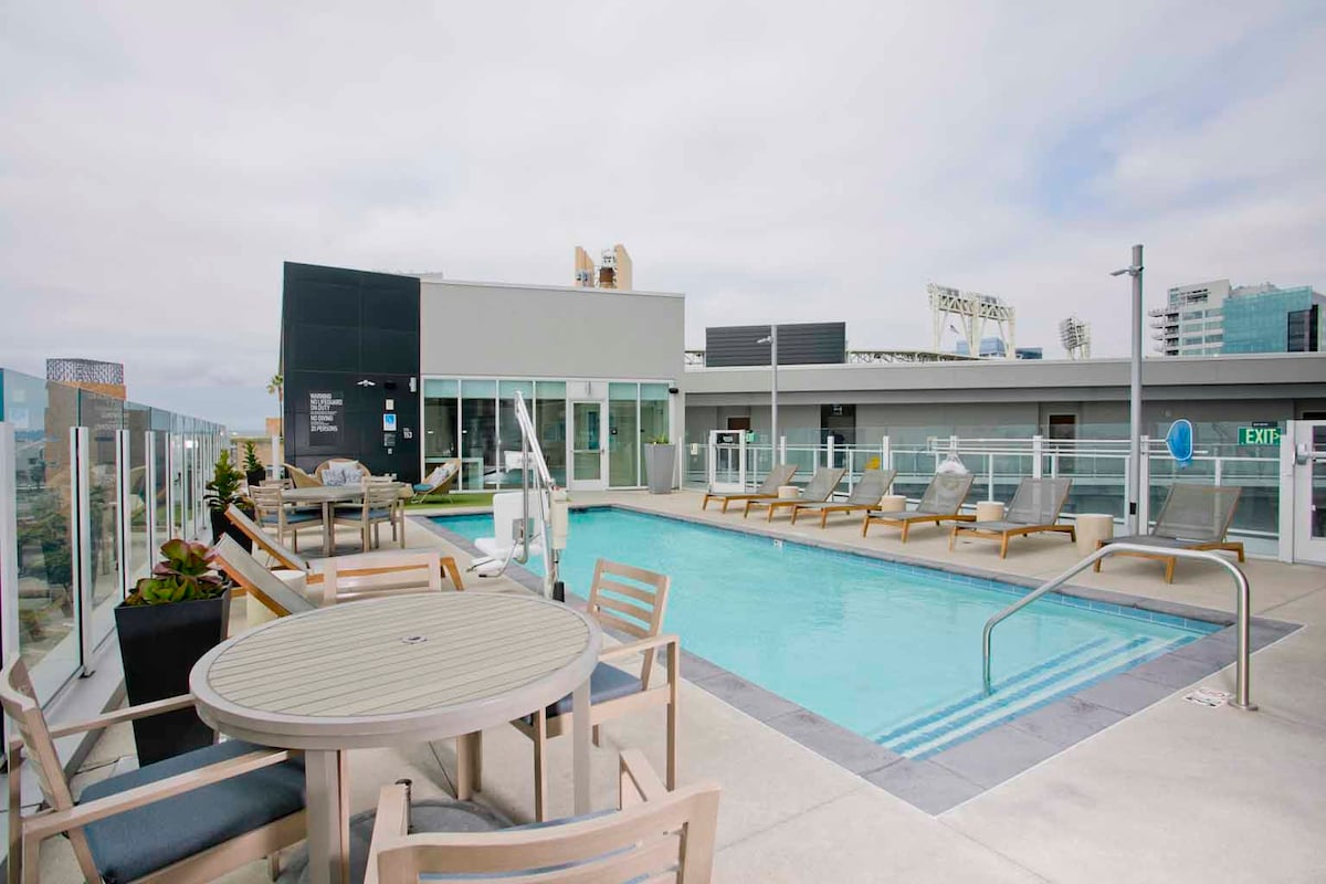 An outdoor pool area is featured, surrounded by lounge chairs positioned against a backdrop of city buildings. A round table is present near the pool, providing a space for gathering. The area is partially enclosed with glass railings, ensuring unobstructed views.