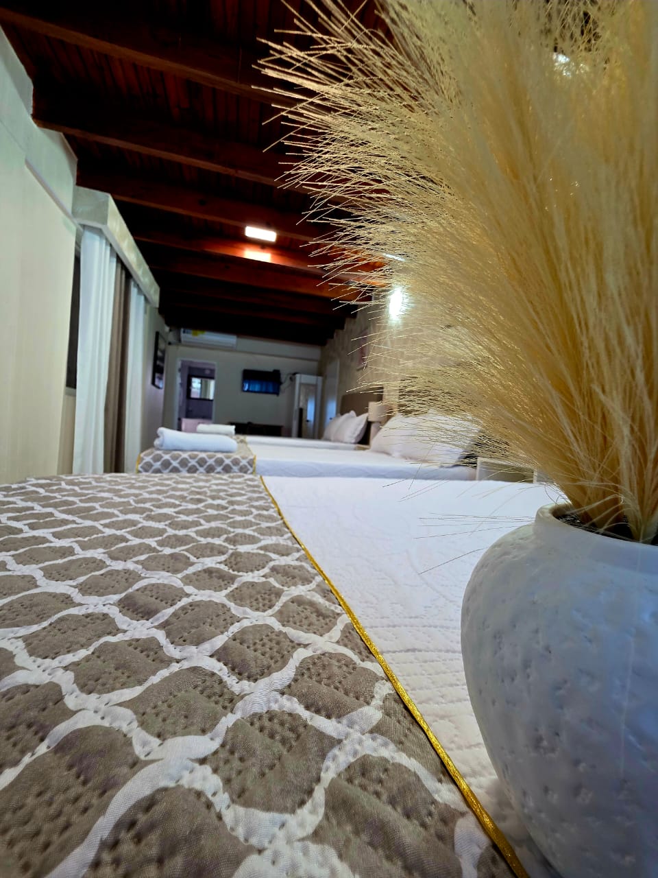 A spacious sleeping area is highlighted by a decorative vase featuring dried plants. The bedding is adorned with a textured coverlet and a neutral palette. Exposed wooden beams are visible on the ceiling, adding natural warmth to the room.