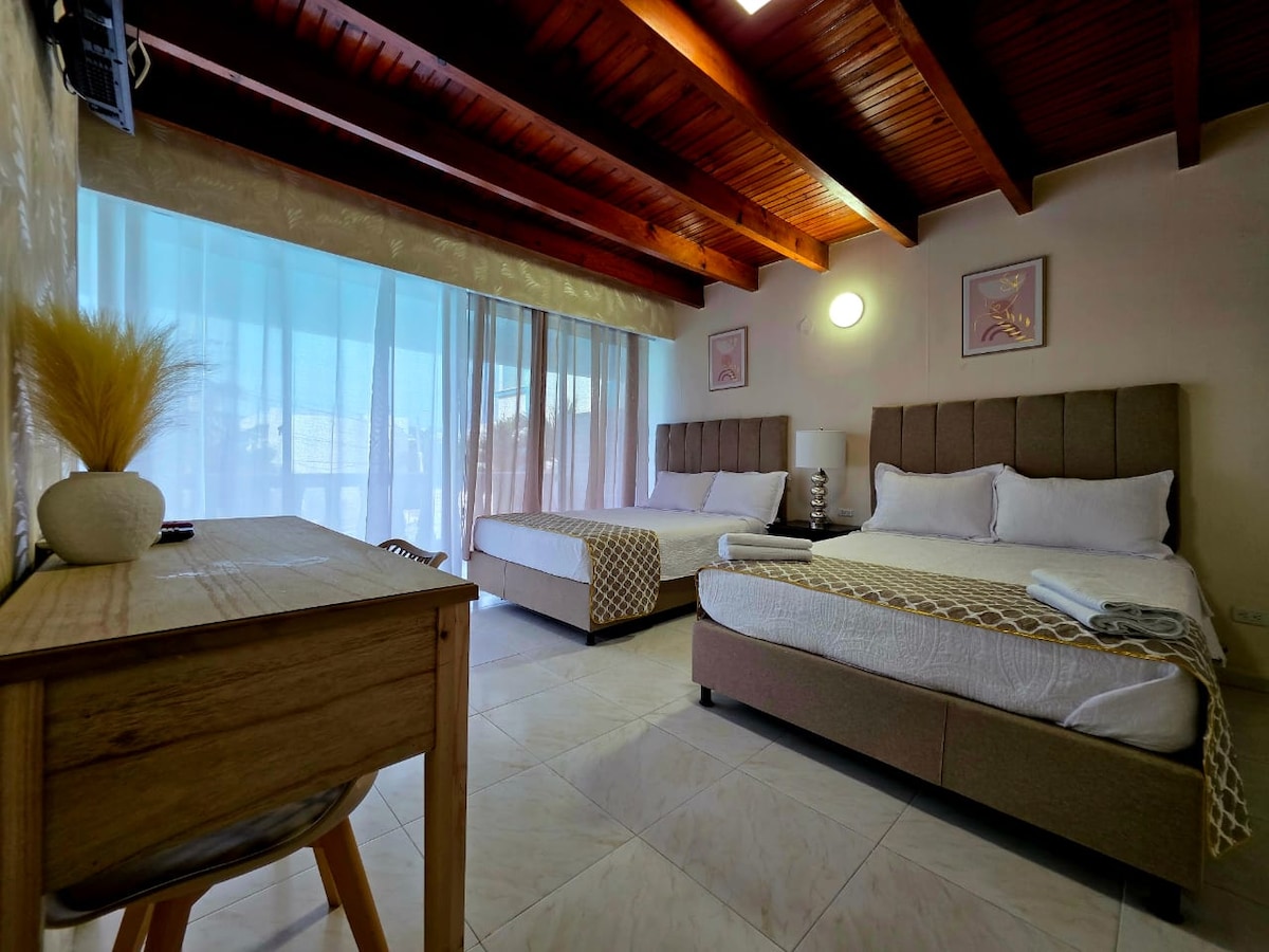 A spacious room features two beds with neutral-colored linens, complemented by light wooden furnishings. Large windows allow natural light to fill the space, while sheer curtains provide a soft filter. A minimalist desk is placed near the window, enhancing the room's functionality.