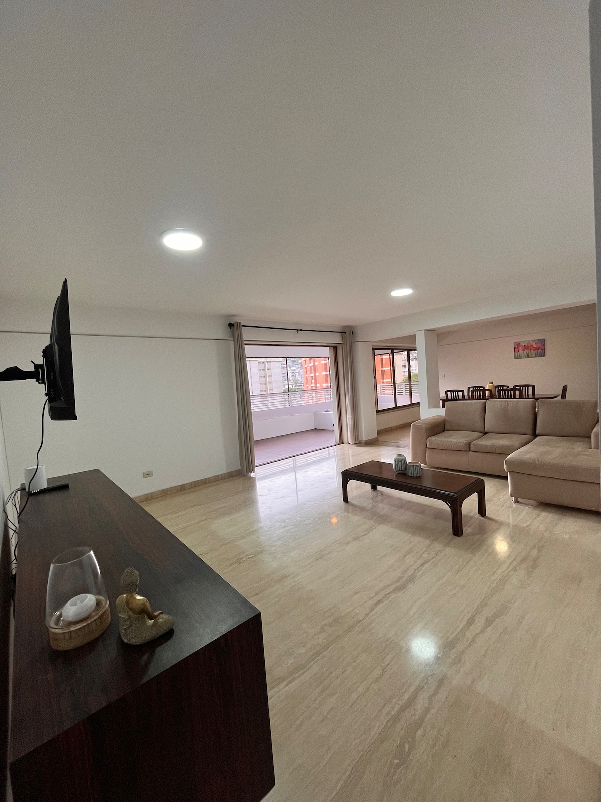 A spacious living area is presented with a light-colored sofa facing a mounted television. A wooden coffee table is centered, and large windows provide natural light. A dining area with a table can be seen in the background, alongside an open terrace access.