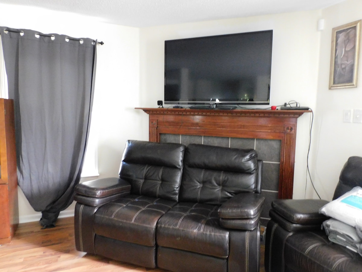 A spacious living area is showcased with a large flat-screen television mounted above a wooden fireplace. Two black leather reclining chairs are positioned for comfort, with a partially drawn dark curtain allowing natural light to filter in. A framed piece of art adorns the wall.