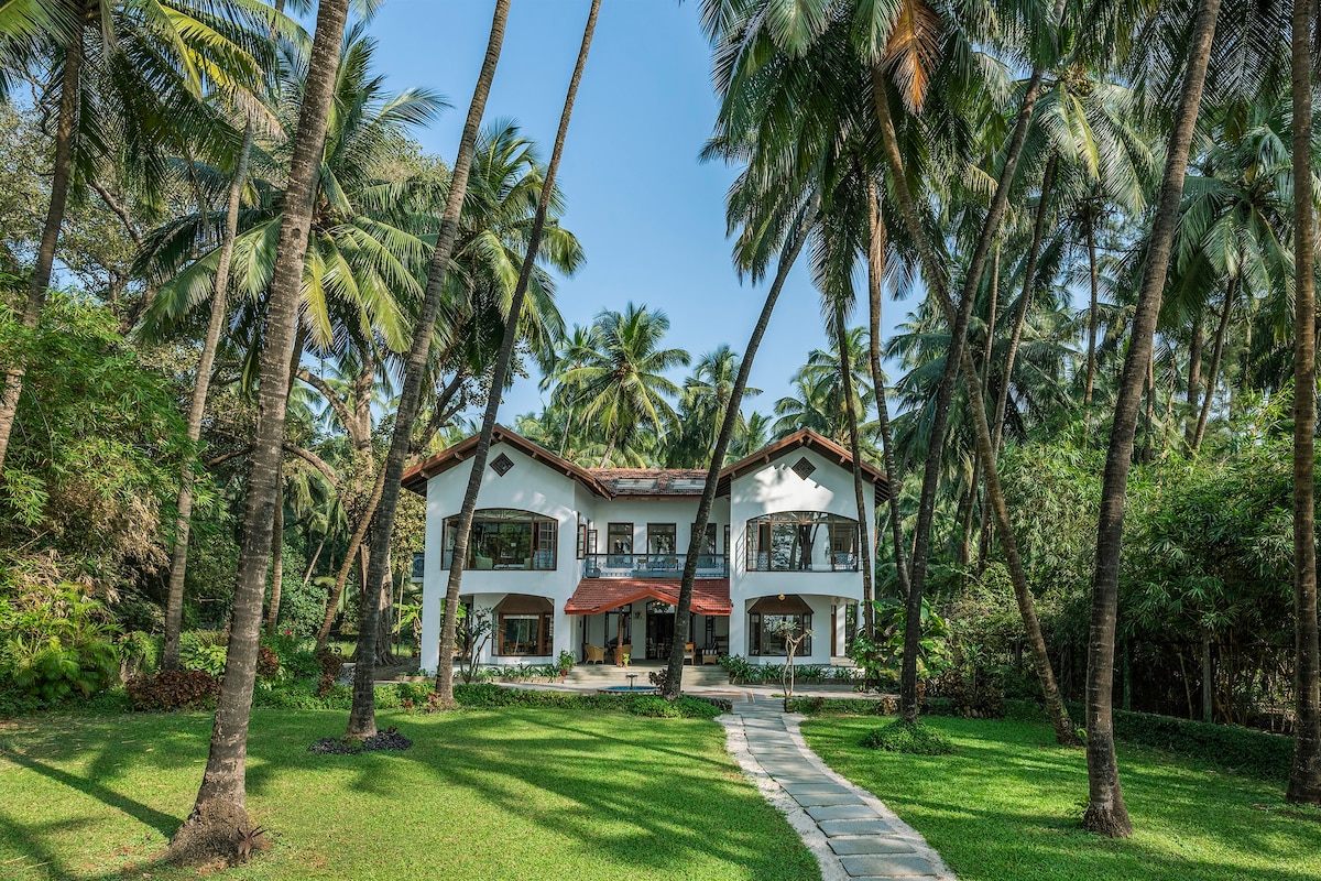 A charming villa stands surrounded by lush, green coconut groves. A stone pathway leads to the entrance, while tall palm trees frame the property under a clear blue sky. The villa features multiple arched windows and a sloping roof, accentuating its inviting architecture.