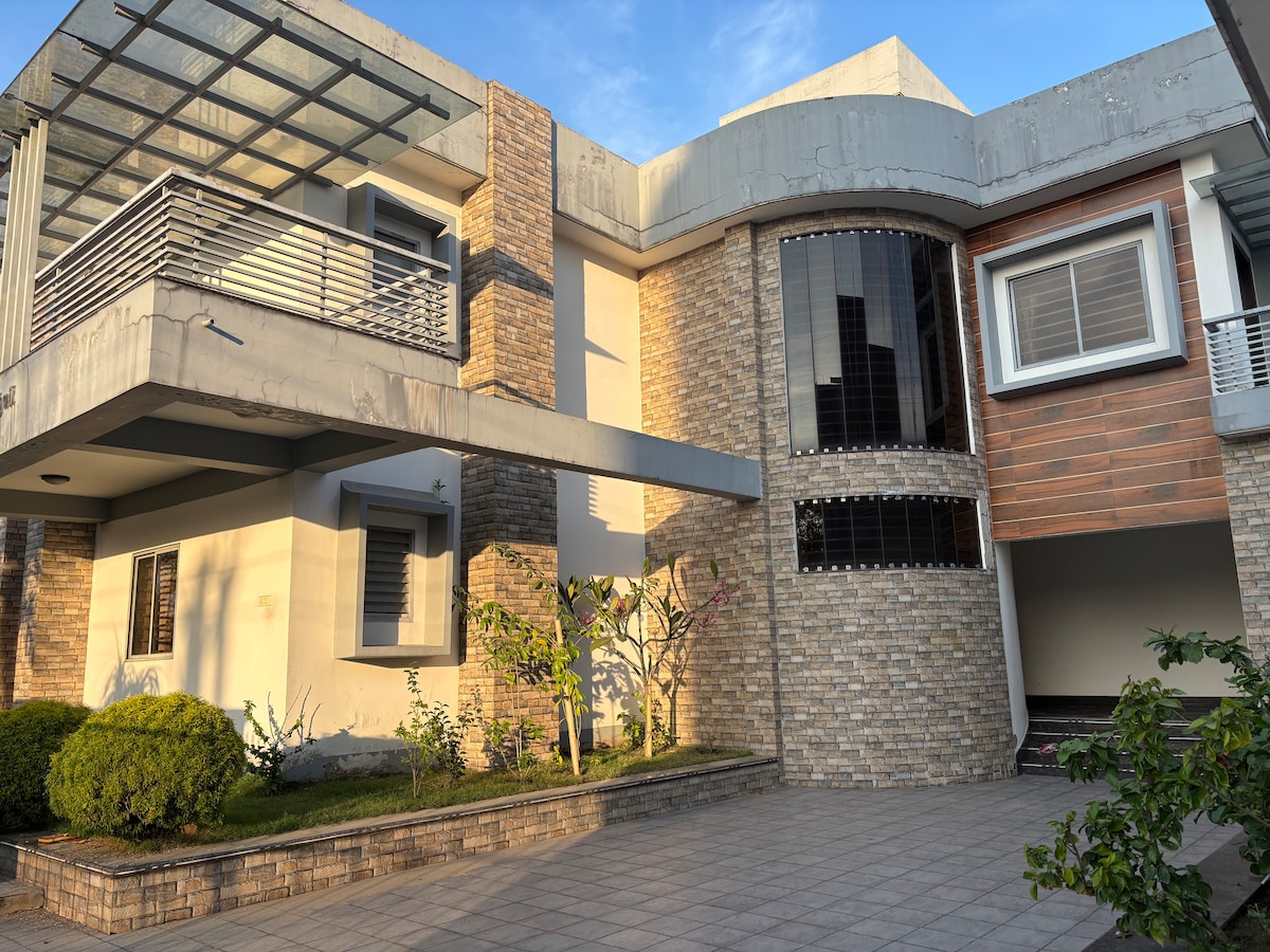 The exterior of the property is showcased, featuring a modern architectural design with a combination of stone and wood materials. A spacious driveway is visible, bordered by well-maintained greenery and minimal landscaping. Natural light enhances the building's clean lines and contemporary look.