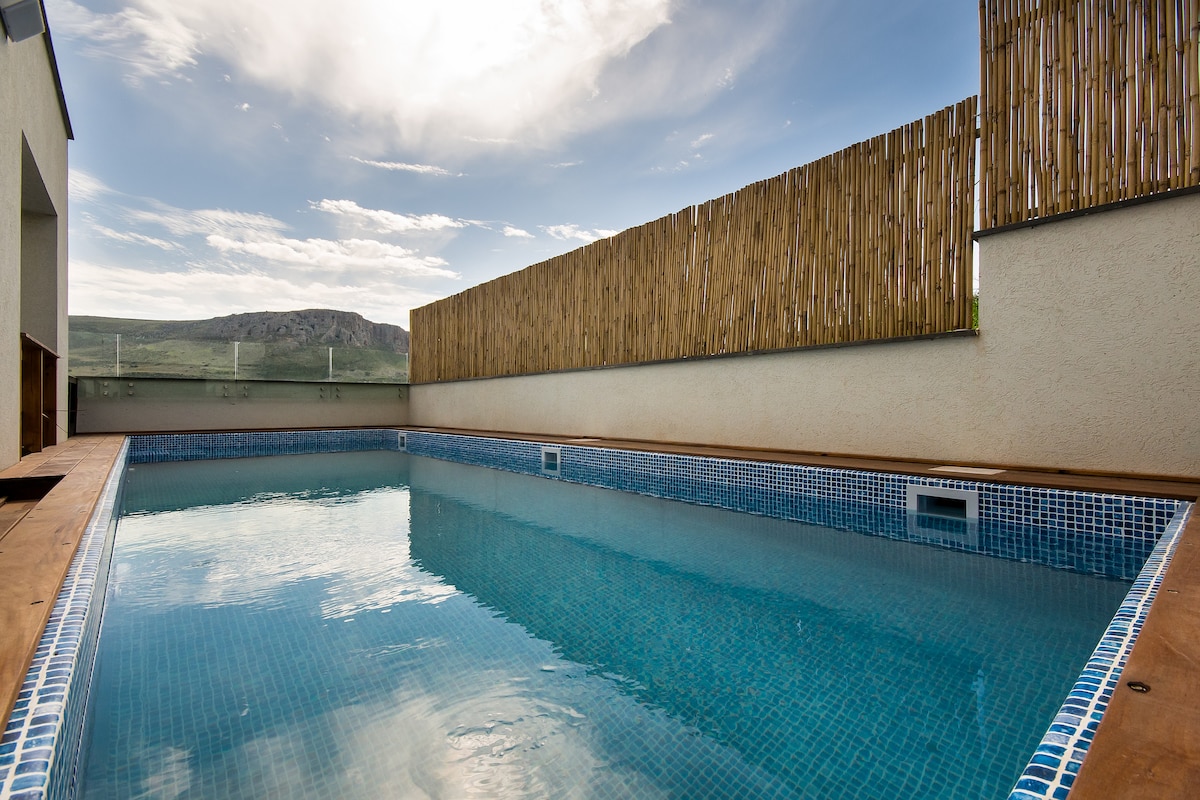 A private swimming pool is surrounded by wooden decking and a tall bamboo privacy fence. Clear water reflects the sky, showcasing a serene atmosphere. The pool's edge is lined with blue mosaic tiles, contributing to an inviting space for relaxation.