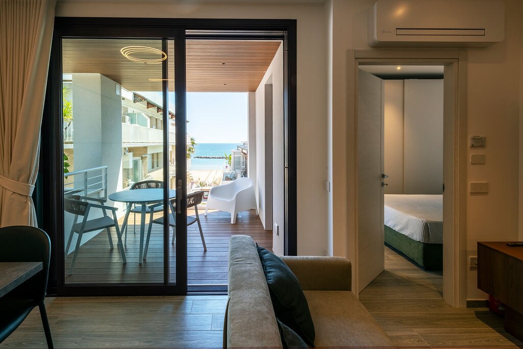 A spacious living area is viewed through sliding glass doors leading to a terrace with a seaside backdrop. Adjacent rooms are visible, including a bedroom with a large bed. Natural light illuminates the space, enhancing the modern design and open layout.
