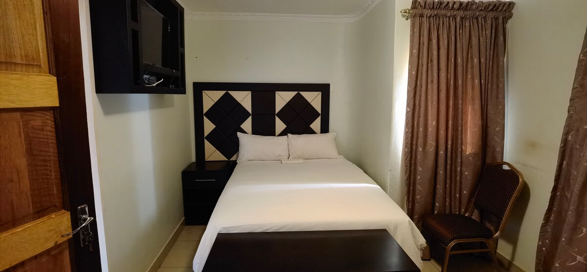 A compact sleeping area features a spacious bed with neutral bedding, complemented by geometric headboard design. A small wooden table is positioned in front of the bed. Natural light filters through sheer curtains, and a wall-mounted TV is visible above a dark bedside table.