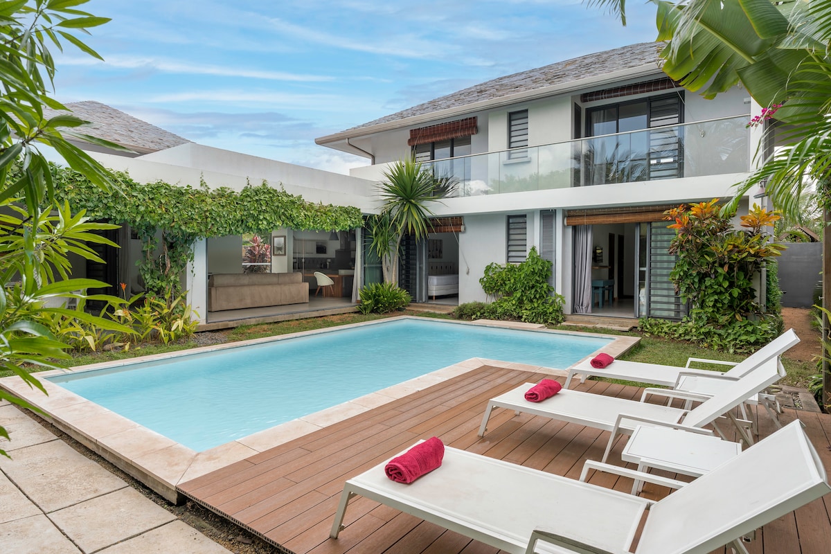A private villa is depicted with a clear swimming pool surrounded by a wooden deck. Sun loungers, adorned with rolled towels, are positioned alongside the pool. Lush greenery and vibrant plants provide a tranquil atmosphere, and large windows reveal a view of the interior living space.