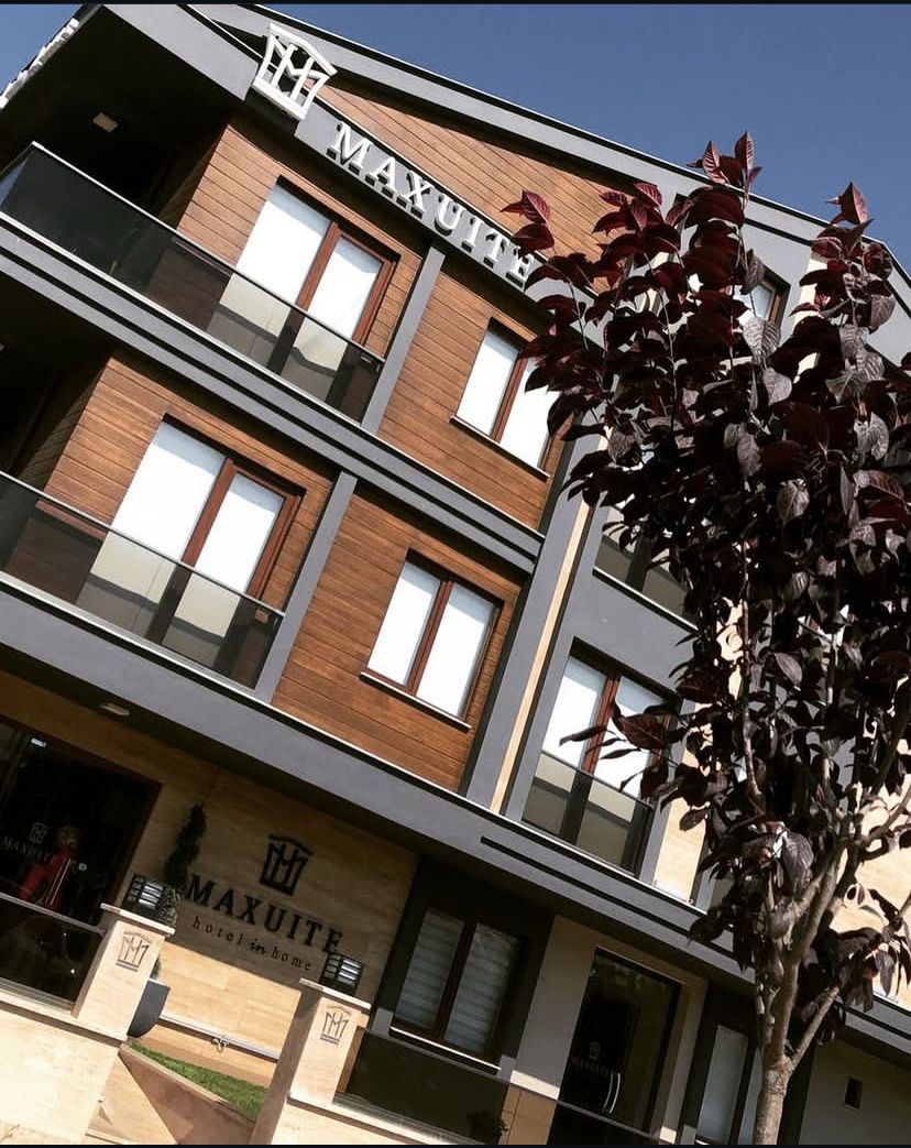 The exterior of Maxuite Hotel showcases a modern building with a combination of wood and grey facades. Several balconies can be seen on the upper floors, while a decorative tree is positioned prominently in the foreground.