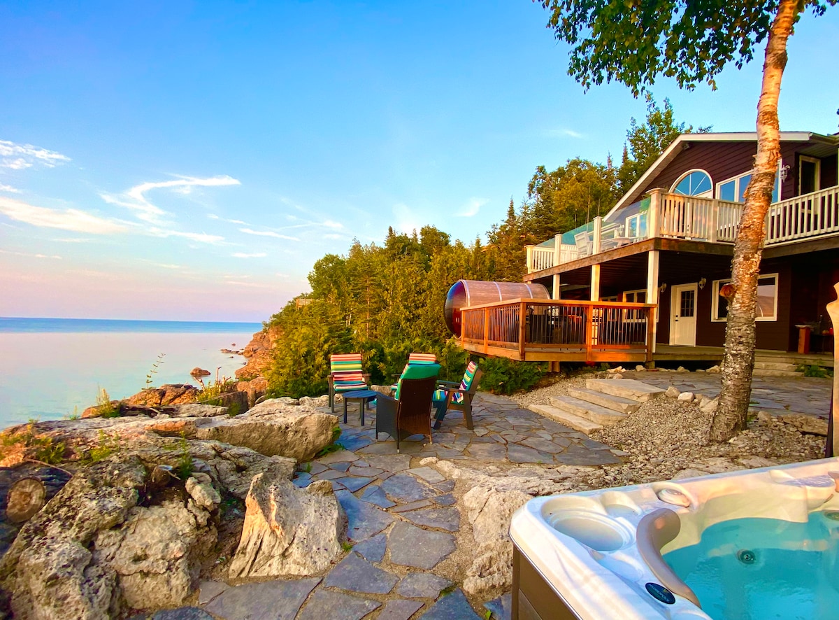 A scenic outdoor area offers breathtaking views of the calm waters and surrounding greenery. A hot tub sits beside a stone patio, equipped with comfortable seating, while a deck extends from the bungalow, providing a relaxing space to enjoy the serene environment.
