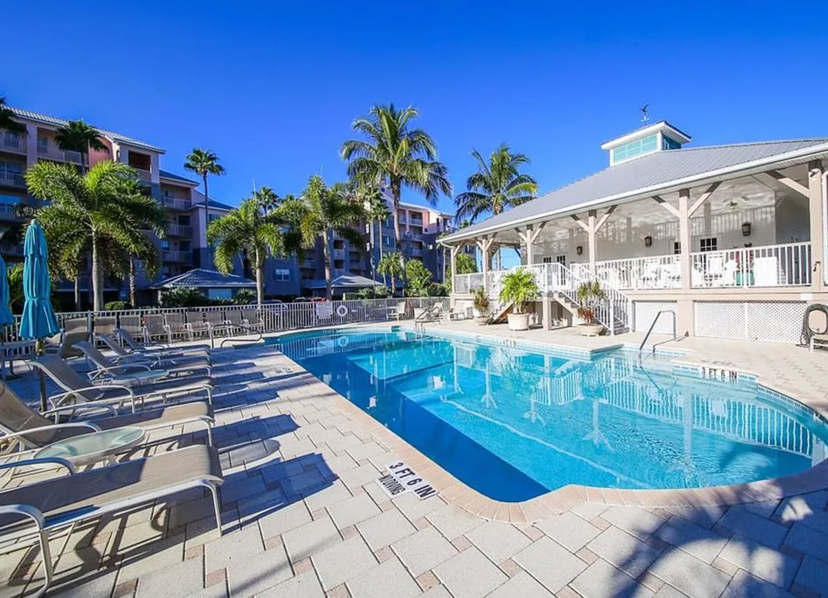A serene pool area is surrounded by lush palm trees and comfortable lounge chairs. The shimmering water is complemented by a covered gazebo, providing shaded seating and relaxation options. Bright blue skies enhance the inviting atmosphere of this outdoor space.