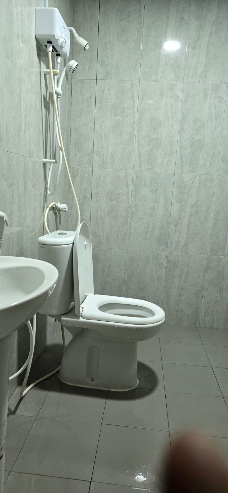 A clean bathroom is depicted with gray tiled walls and flooring. A white toilet and a small basin are positioned against the wall. A showerhead is mounted above the toilet, and a white hot water system is installed next to the shower.
