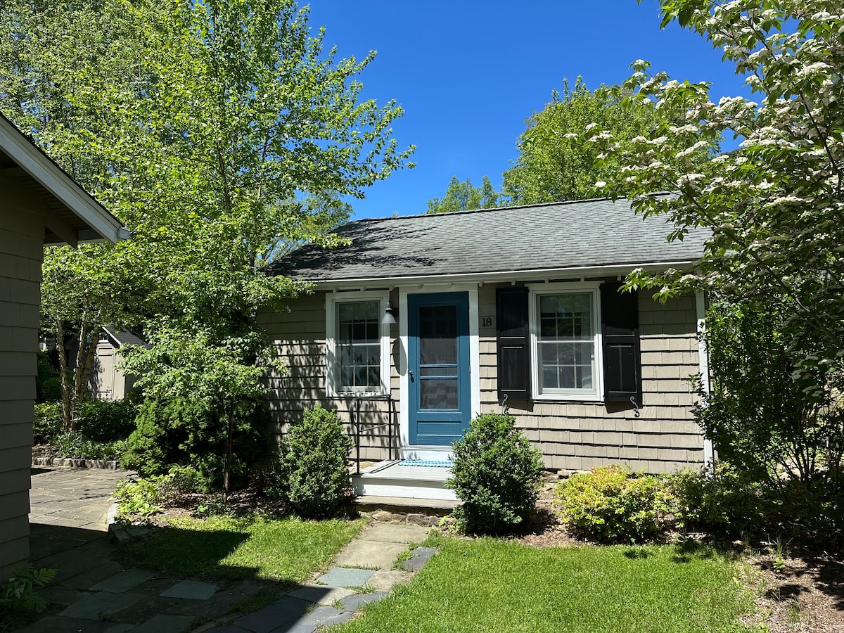 The cozy cottage is framed by lush green shrubs and trees, with a welcoming blue front door. A stone pathway leads to the entrance, surrounded by a well-maintained garden. Bright blue skies enhance the serene outdoor environment.