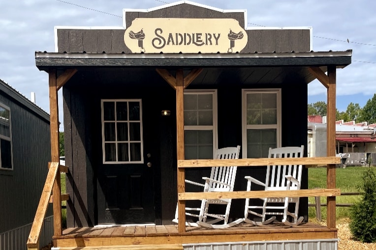 The exterior of a cabin, featuring a porch with two rocking chairs. A sign labeled 'SADDLERY' is mounted above the entrance. The cabin has a dark exterior with a single window beside the wooden door, surrounded by greenery.