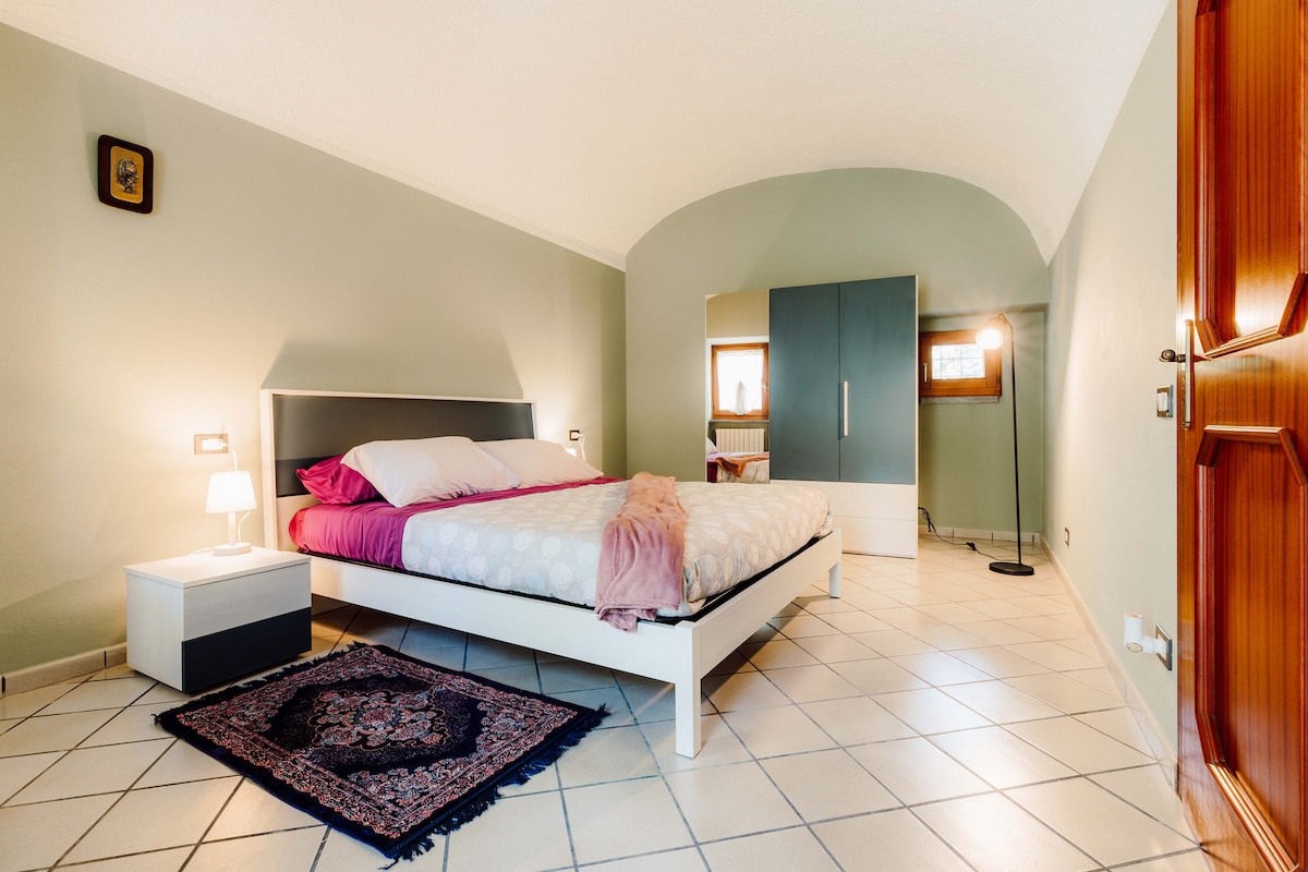 A spacious bedroom features a comfortable bed with light bedding and accent pillows. A nightstand with a lamp is positioned beside the bed, while a wardrobe and a decorative rug add functionality and warmth to the room. Natural light enters through a window, enhancing the serene atmosphere.