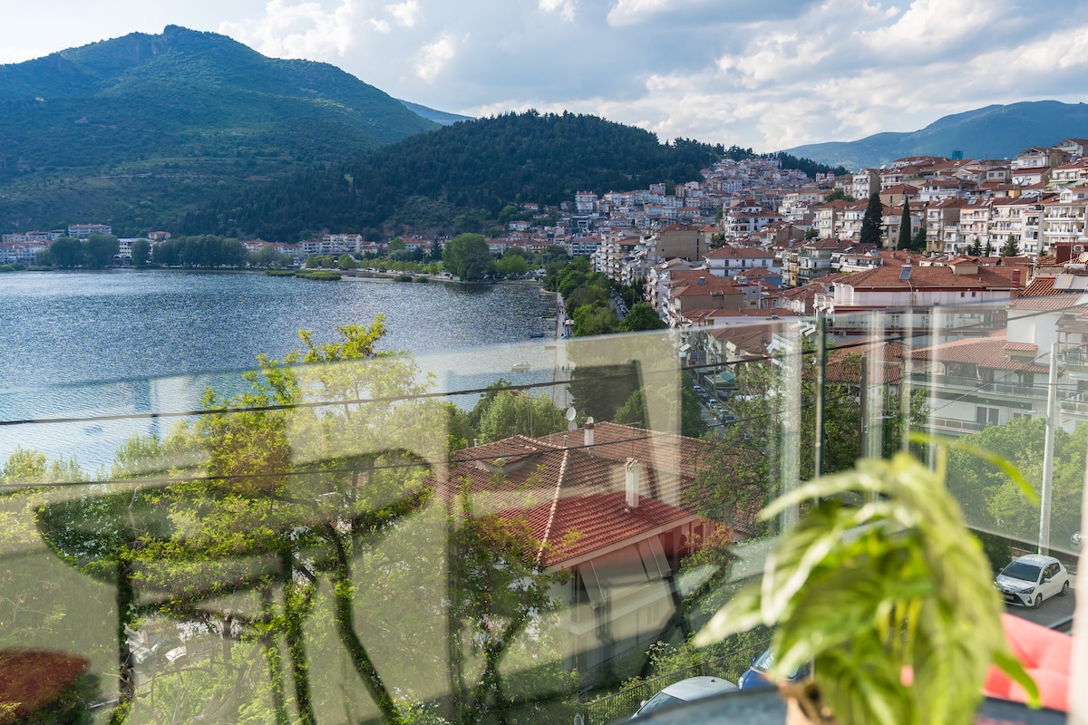 A panoramic view of a lakeside town is featured, with lush mountains rising behind. The image captures a blend of rooftop tiles and greenery, reflecting the serene water and surrounding landscape. Clear skies allow natural light to illuminate the scene, enhancing the vibrant colors.