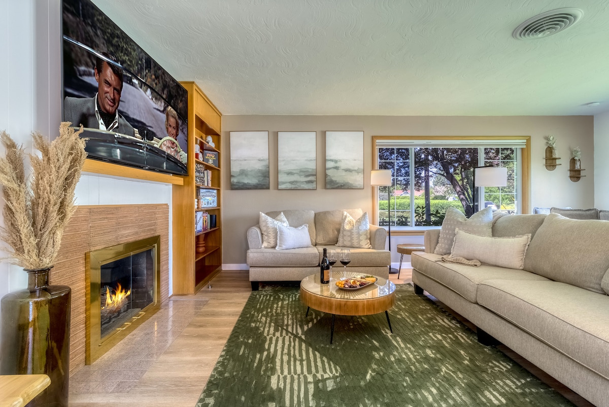 A welcoming living area is captured, featuring a cozy arrangement of light-colored sofas positioned around a circular coffee table. A media unit holds a large television above a modern fireplace. Natural light floods the space from a wide window overlooking greenery.
