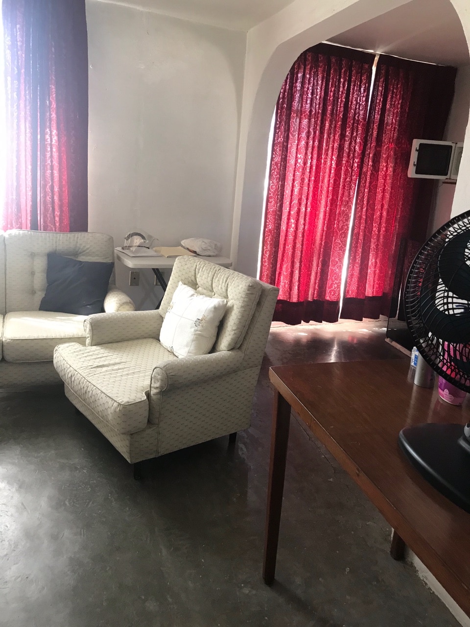 A bright living space features two light-colored armchairs with decorative cushions. Dark red curtains frame a window, allowing soft light to filter in. A wooden table with a fan is positioned nearby, complementing the simple and serene design of the area.