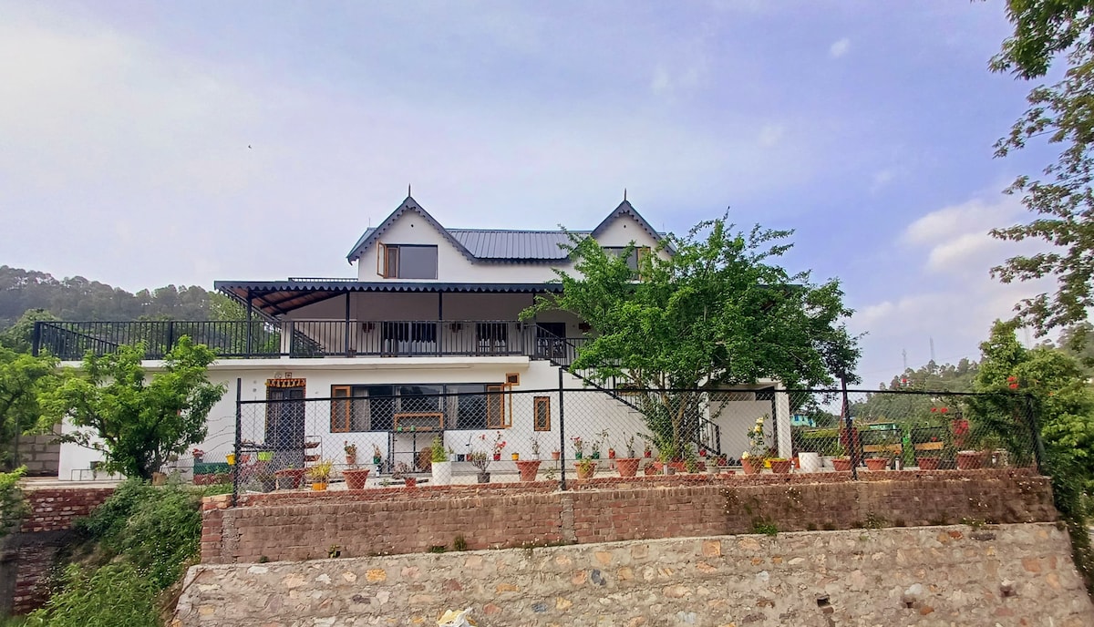 The homestay is showcased from a distance, featuring a two-story structure with a sloped roof and multiple gabled peaks. Surrounding gardens with potted plants are visible. A protective fence and stone wall encase the property, set against a backdrop of lush greenery and a clear sky.