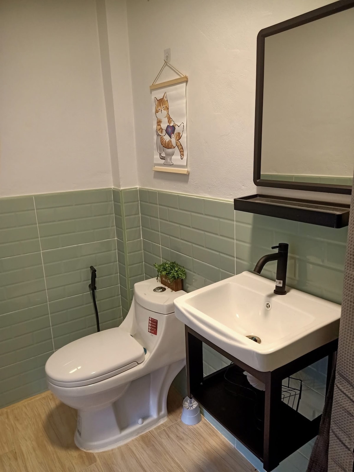 A compact bathroom features mint green tiled walls and light wood flooring. A white toilet is positioned next to a modern black sink with a sleek faucet. A wall-mounted mirror and a small decorative plant add subtle accents to the space.