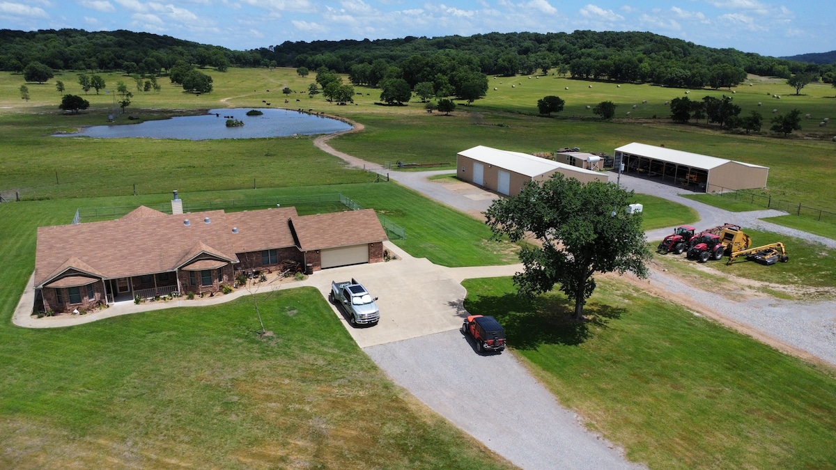 An aerial view of the sprawling ranch property is displayed, featuring a single-story house with a tan roof and ample parking space. Adjacent structures, including a horse barn and storage facilities, are visible. A pond and green pastures dominate the landscape, dotted with trees.
