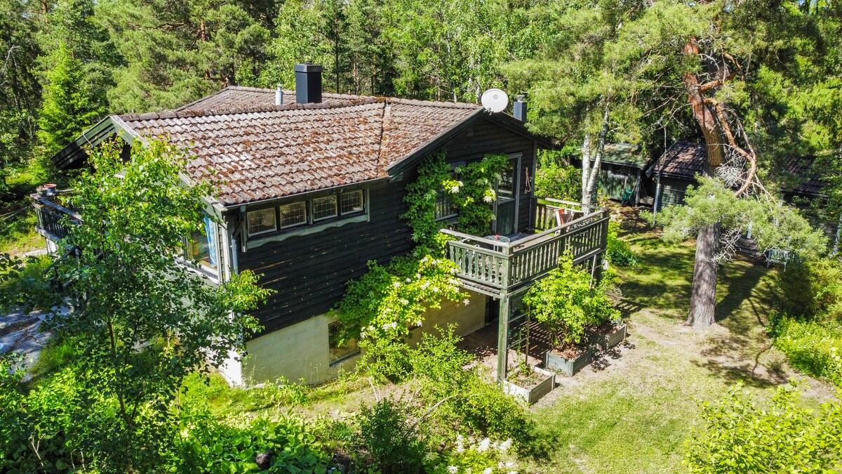 A traditional house from the 70s is surrounded by lush greenery and trees. The exterior features a blend of dark wood with a sloped roof, complemented by a spacious balcony. A nearby patio is visible, showcasing an inviting outdoor space.