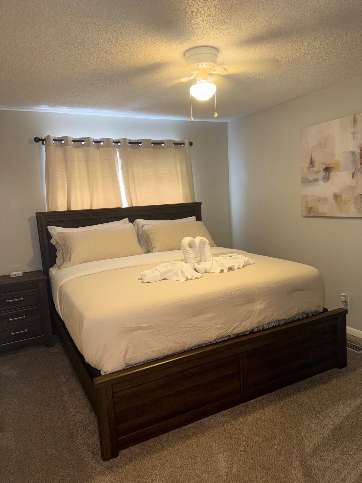 A well-appointed bedroom features a spacious bed with light linens arranged neatly. A ceiling fan is positioned above, providing a gentle breeze. Natural light filters through light curtains, illuminating a subtle wall art piece to the side. A bedside table is located next to the bed.