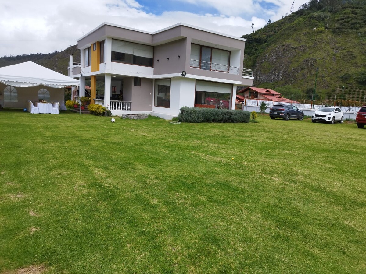 A modern two-story house is depicted, featuring large windows and a balcony. A spacious lawn surrounds the property, with neatly trimmed grass. In the foreground, a white tent is visible, accompanied by a set of tables, suggesting an area for outdoor gatherings. The landscape showcases gentle hills in the background.