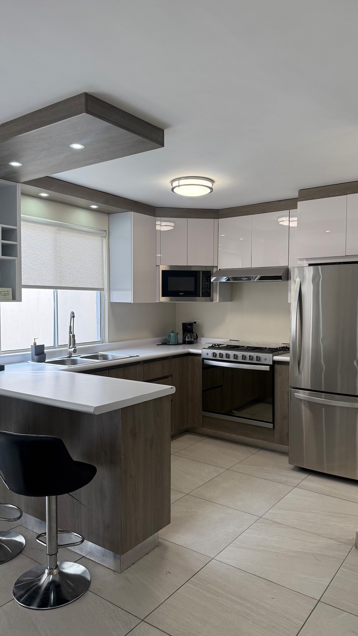 A modern kitchen showcases sleek cabinetry in a combination of light and dark wood finishes. Stainless steel appliances, including a refrigerator and oven, are visible. A small island with high-backed stools provides a space for casual dining or preparation.