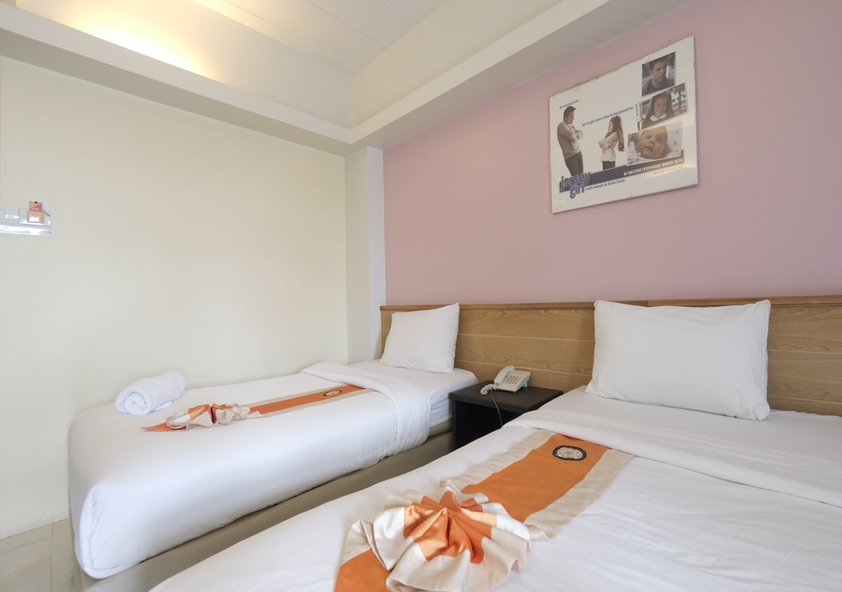 A well-lit sleeping area features two spacious beds adorned with soft, white linens and decorative orange accents. A bedside table and a telephone are positioned beside one bed, while a wall-mounted shelf shows a few neatly arranged items. A light pink wall complements the warm tones.