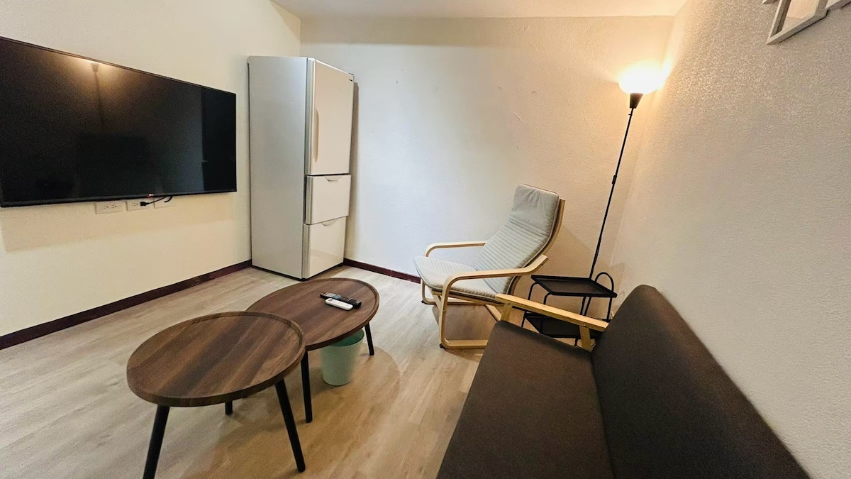 A living space features a brown sofa and a light-colored armchair, complemented by two round coffee tables. A large television is mounted on the wall opposite, with a refrigerator placed to the side. Soft lighting from a floor lamp adds to the ambiance.