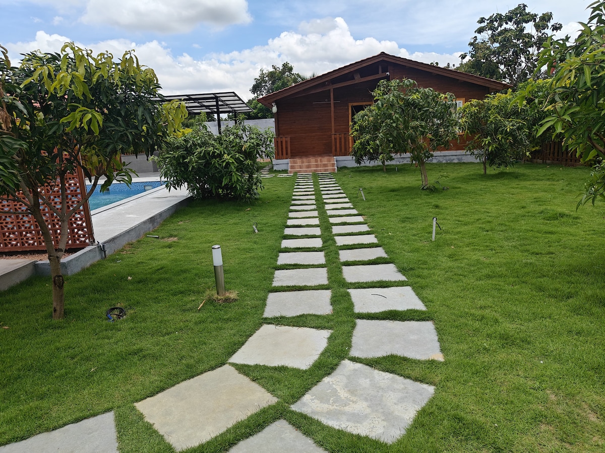A well-maintained garden path of stone slabs leads towards a wooden cabin surrounded by lush greenery. The landscape features manicured grass and several trees, creating a serene outdoor environment. A pool area is visible in the background, enhancing the tranquil setting.