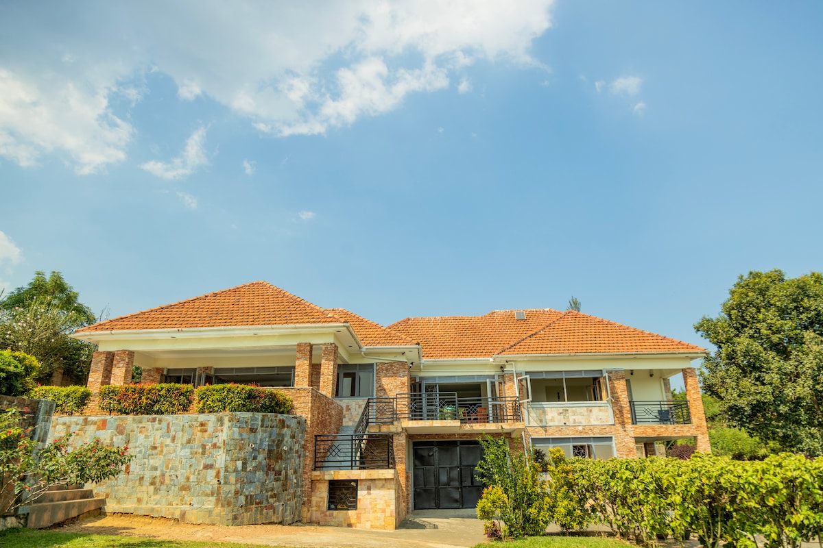 A spacious villa is depicted against a vibrant blue sky, featuring a combination of stone and brick exterior. The well-manicured grounds are visible, with green foliage accentuating the entrance steps and surrounding the home's entrance.