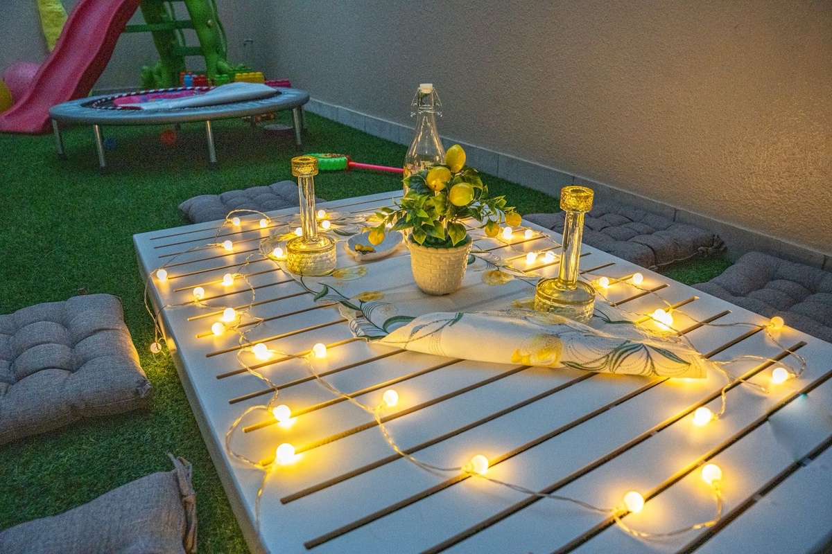 A neatly arranged outdoor table is set with decorative floral accents and illuminated by string lights, creating a warm atmosphere. Soft cushions are positioned on the grass, alongside a trampoline and play equipment, indicating a family-friendly environment.