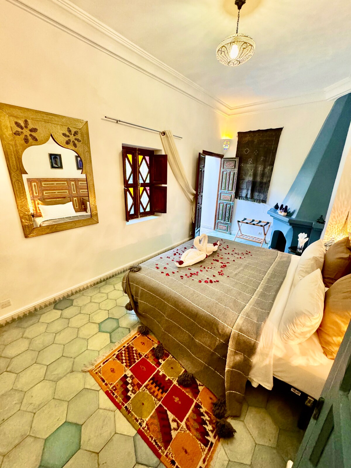A serene bedroom features a spacious bed adorned with a soft grey blanket and decorative pillows. A mirror reflects the room's design, while light filters through window shutters. A hint of decorative petals adds a touch of elegance to the inviting atmosphere.