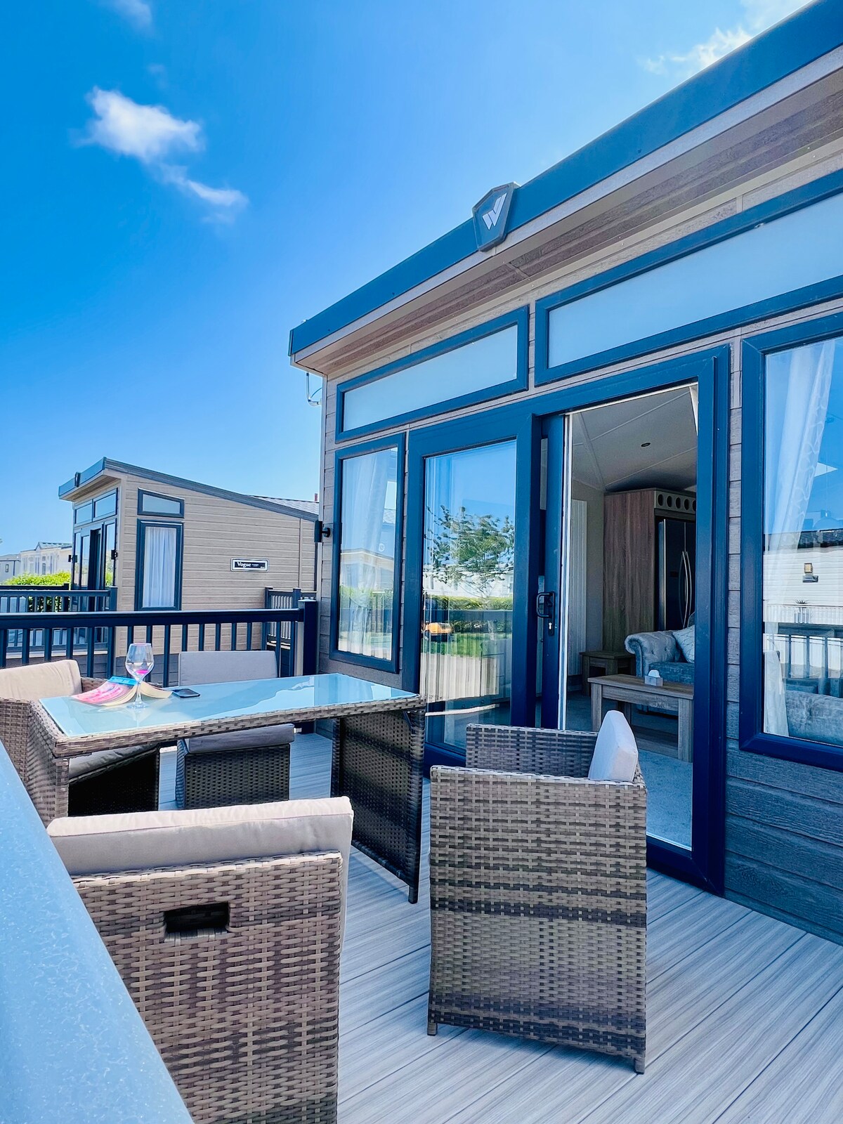 A balcony area is depicted, featuring a glass-topped table accompanied by two woven chairs. Large glass doors provide entry to the interior, framed by a sleek, modern exterior. The space is illuminated by natural light, with blue skies visible in the background.