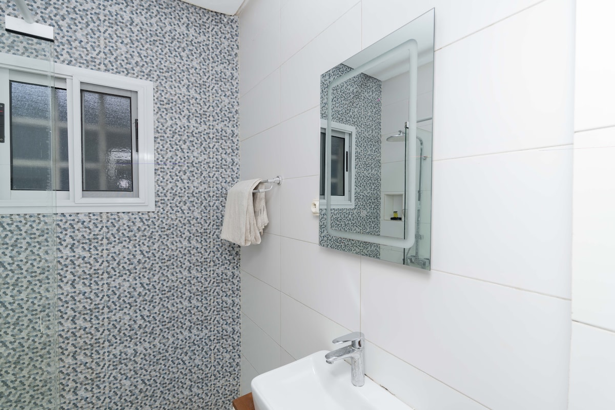 A bright bathroom features a modern sink with a sleek faucet and a wall-mounted mirror. A textured wall in subtle blue tones adds interest, while a small window allows natural light. A towel hangs on a rack, enhancing the practical elements of the space.