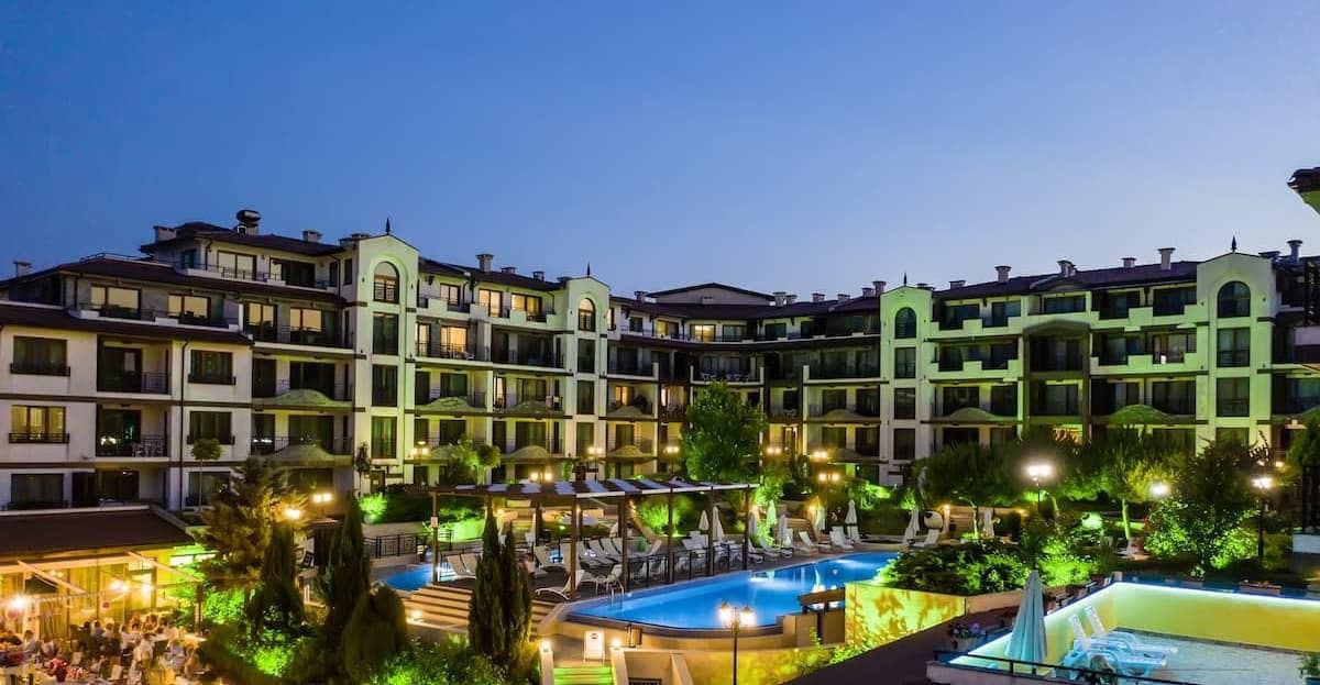 The image presents a modern residential complex at dusk, featuring multiple levels illuminated by soft lighting. A spacious outdoor pool is visible, surrounded by lounge chairs and lush greenery, while dining areas are apparent nearby, enhancing the inviting atmosphere of the complex.