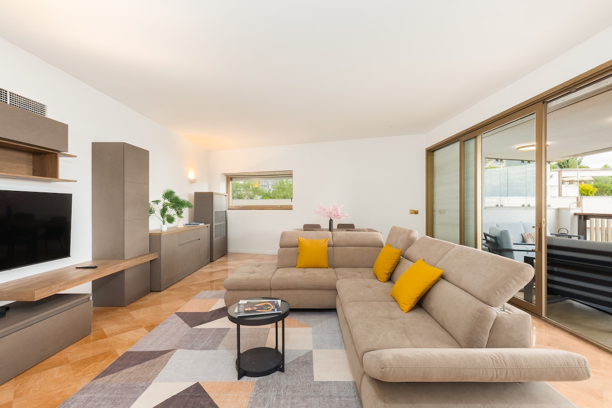 A spacious living area is displayed, featuring a soft, neutral-colored sectional sofa with bright yellow accent pillows. Large glass doors open to a terrace, allowing natural light to fill the room. A contemporary coffee table sits in front of the sofa, complemented by modern shelving and a television.
