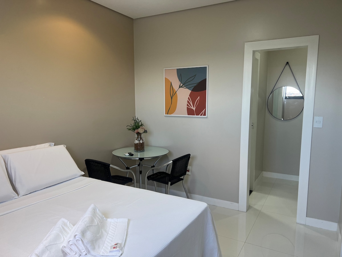 A clean and modern room features a spacious bed with crisp white linens and a small dining area with a glass table and two black chairs. A colorful wall art piece is displayed, and a round mirror hangs near the entrance, reflecting the soft lighting of the room.