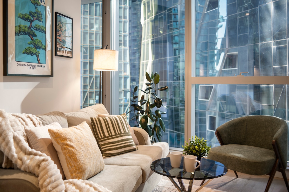A cozy living area features a light-colored sofa adorned with textured cushions and a soft throw. A round coffee table holds two cups and a small plant. Large windows allow natural light to flood the space, revealing reflections of the cityscape outside.
