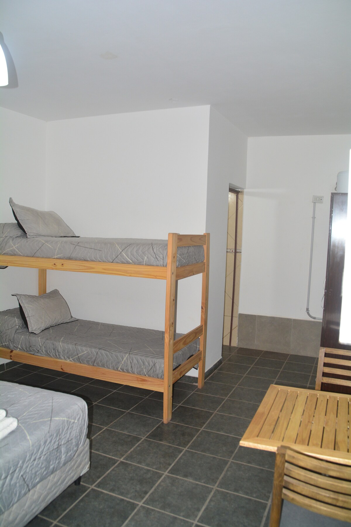 A simple sleeping area features a wooden bunk bed with gray bedding and neatly arranged pillows. A small wooden table is positioned nearby, accompanied by chairs. Neutral walls and tiled flooring create a clean, spacious environment.