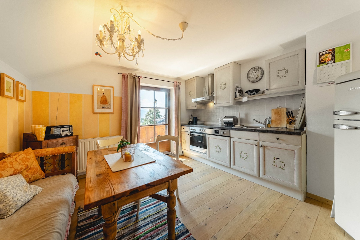 A cozy kitchen area features a rustic wooden table surrounded by comfortable seating. Light wood flooring complements the simple cabinetry and modern appliances. A window allows natural light to flood the space, highlighting the warm tones of the decor and inviting atmosphere.