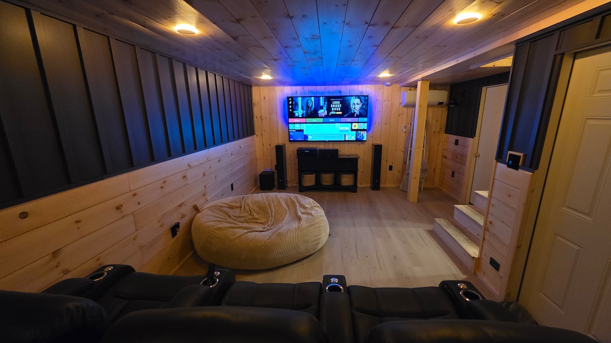 A home theater room features leather reclining seats arranged for comfort, facing a large screen mounted on a wooden wall panel. Soft ambient lighting enhances the cozy atmosphere, while a plush bean bag chair offers additional seating. Steps lead to a well-organized area with media storage.