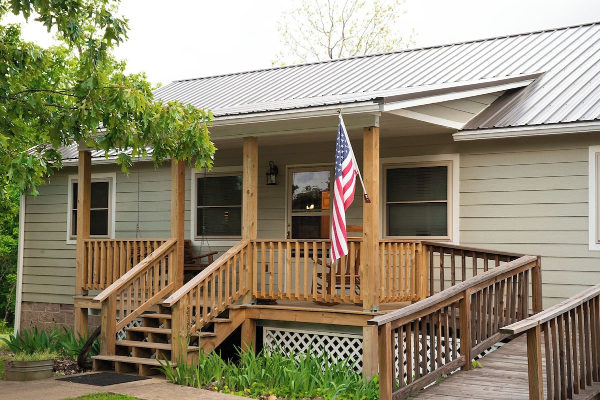 A welcoming front porch features a wooden railing and a set of stairs leading to the entrance of the cabin. An American flag is displayed proudly, and greenery is visible surrounding the area, creating an inviting outdoor space.