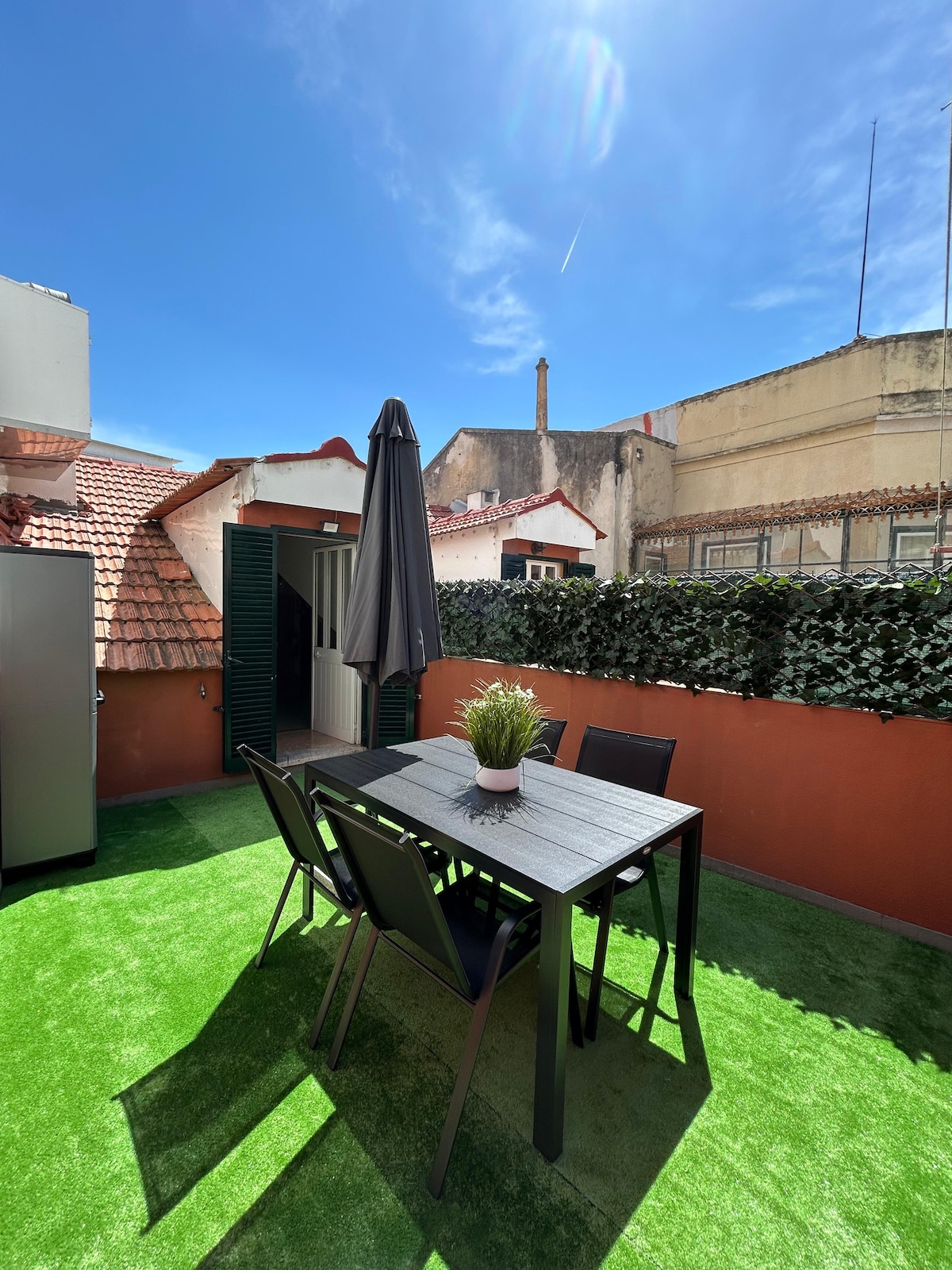 A spacious terrace is presented, featuring a black dining table surrounded by six chairs. A large umbrella provides shade, and a small potted plant adds a touch of greenery. The terrace is set against a backdrop of rooftops and clear blue sky.