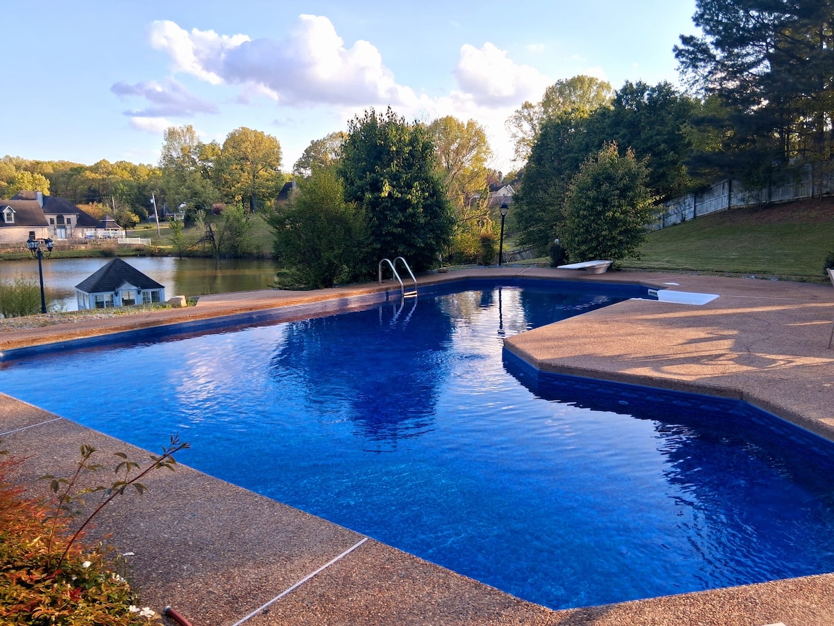A tranquil private pool is surrounded by lush greenery and a landscaped yard. Reflective blue water invites relaxation, while gentle steps lead into the pool. A serene lake is visible in the background, enhancing the peaceful atmosphere of the outdoor space.