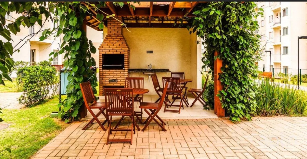 An outdoor seating area features wooden tables and chairs arranged under a covered pergola. Lush green vines adorn the structure, and a built-in barbecue grill is positioned against a brick wall. The surrounding landscape includes manicured grass and shrubs.