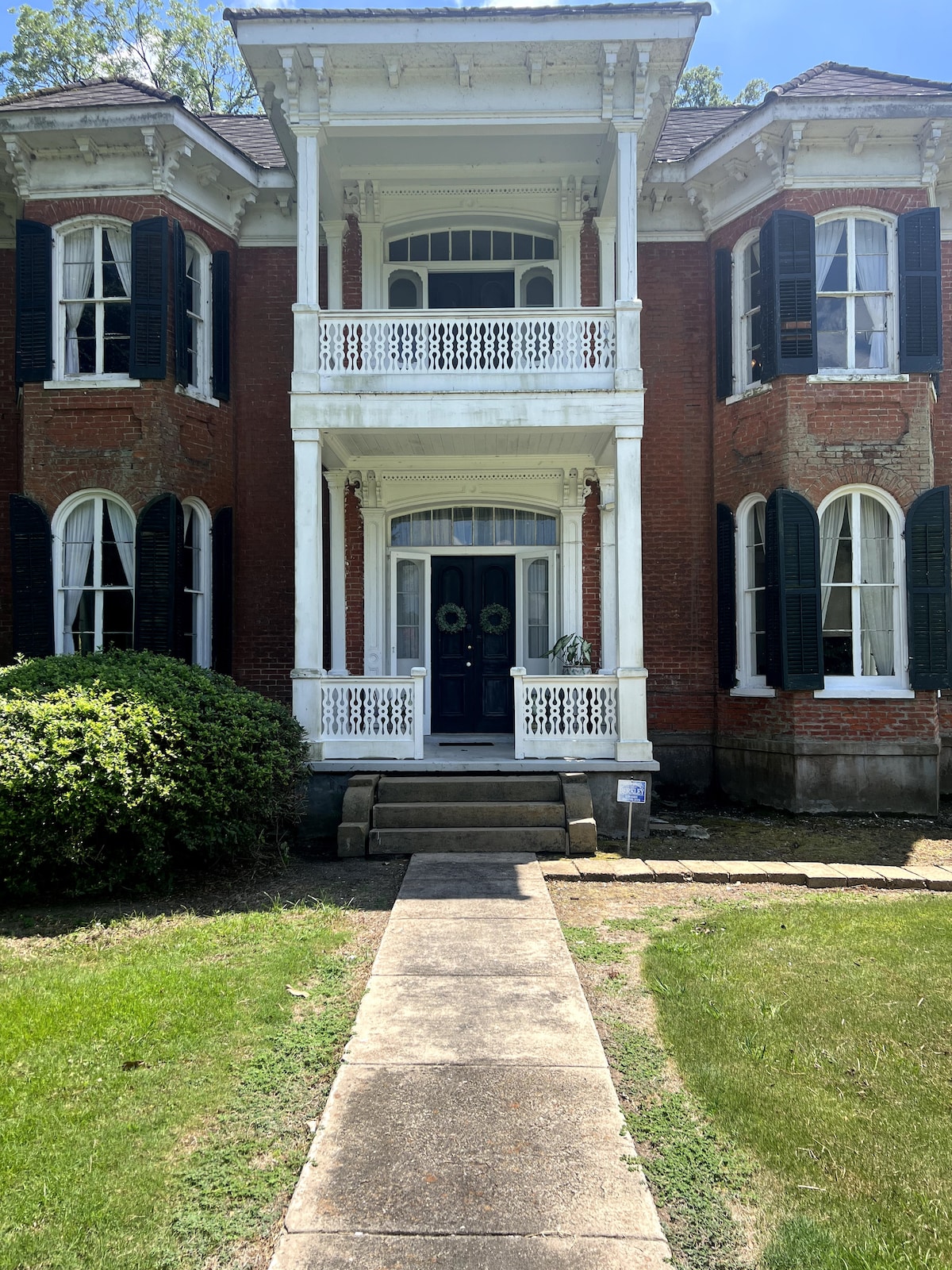 A historic red brick house features a grand entrance with a white balcony and detailed trim. Lush greenery surrounds the path leading to the front door, which is framed by two elegant columns. Dark shutters add contrast to the light facade, enhancing the architectural details.