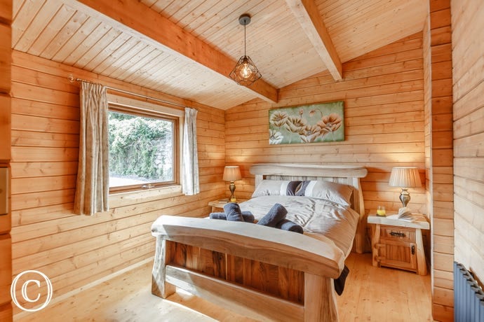 A rustic bedroom features handcrafted wooden paneling and beams, complemented by a large window providing natural light. A spacious bed is adorned with neatly arranged towels, flanked by two bedside lamps. Wall art depicting a floral motif adds a subtle touch of decor.