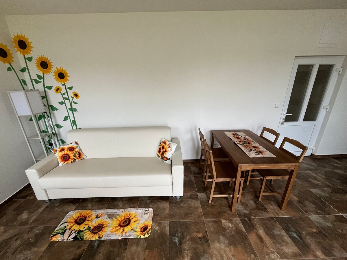 A light-filled living space features a white sofa adorned with floral cushions, complemented by a wooden dining table surrounded by six chairs. Sunflower wall decals add a cheerful touch to the room, while a lamp stands beside the sofa, enhancing the inviting ambiance.